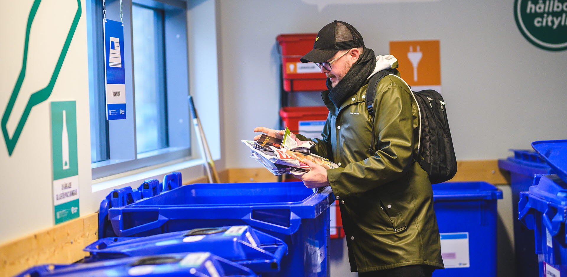 En man står i ett återvinningsrum och håller i en bunt tidningar ovanför ett blått avfallskärl.