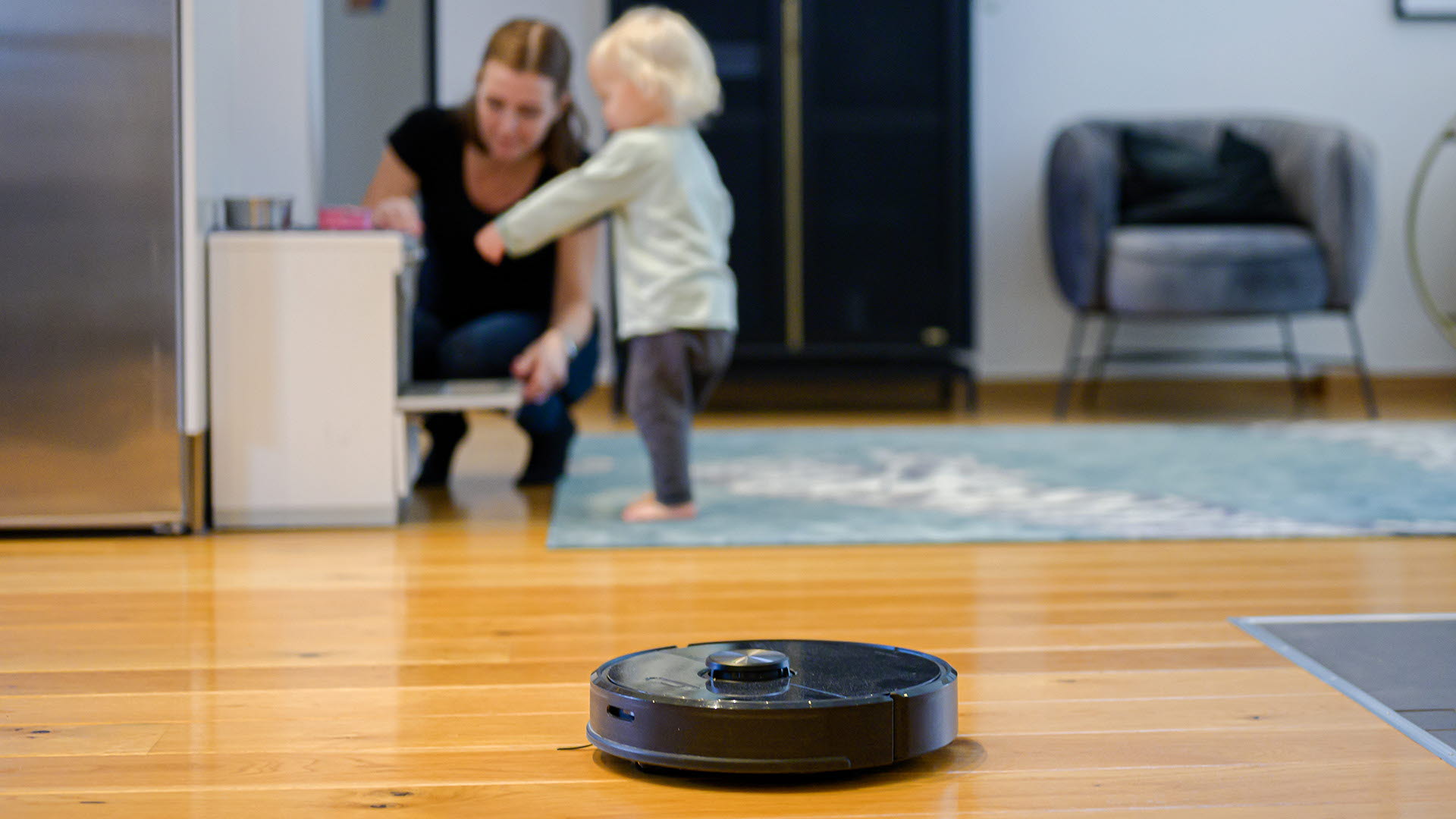 En robotdammsugare dammsuger giolvet medan en mamma och litet barn leker i bakgrunden.